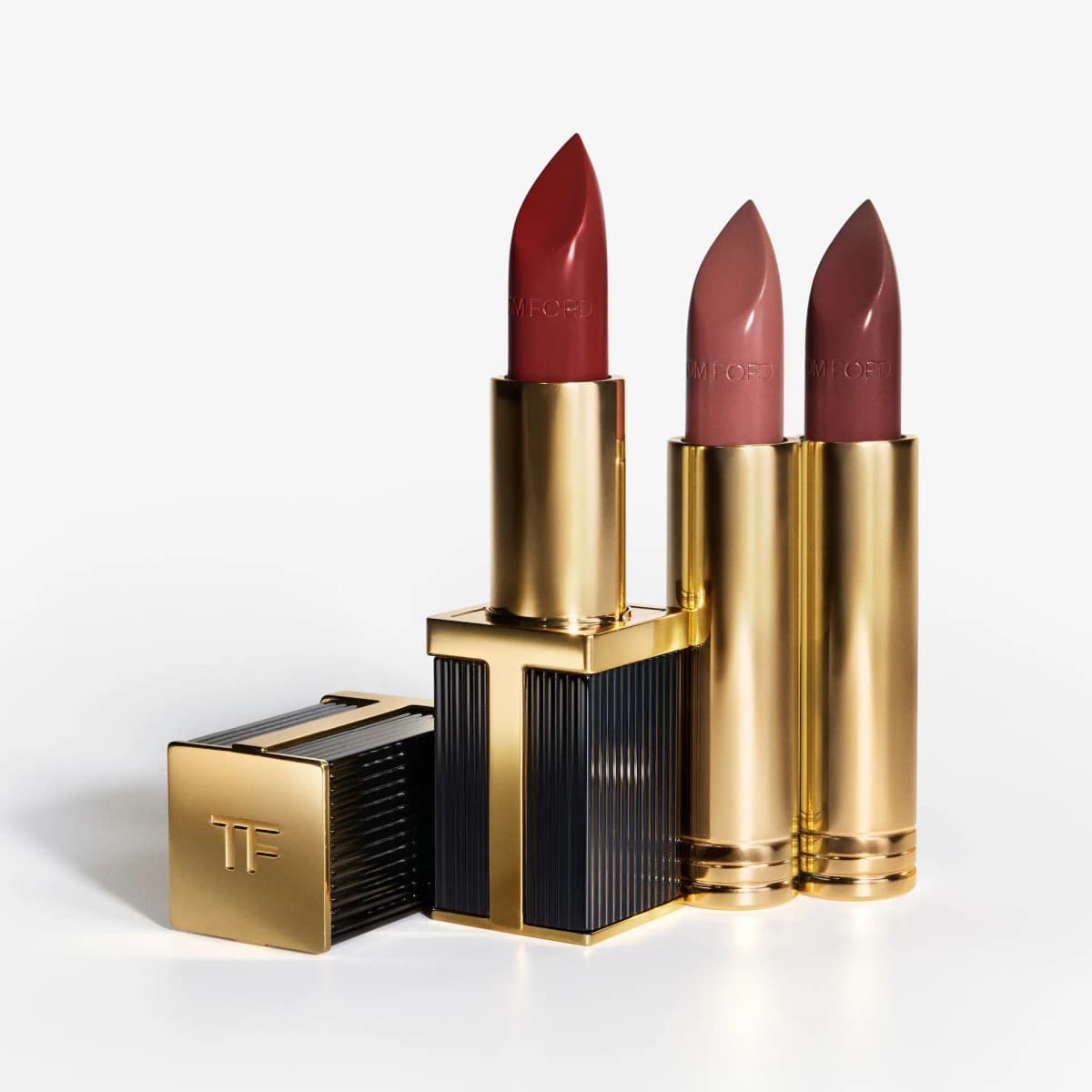 Open black-and-gold ribbed case holding a red lipstick, beside its TF cap and two gold refills in nude and mauve
