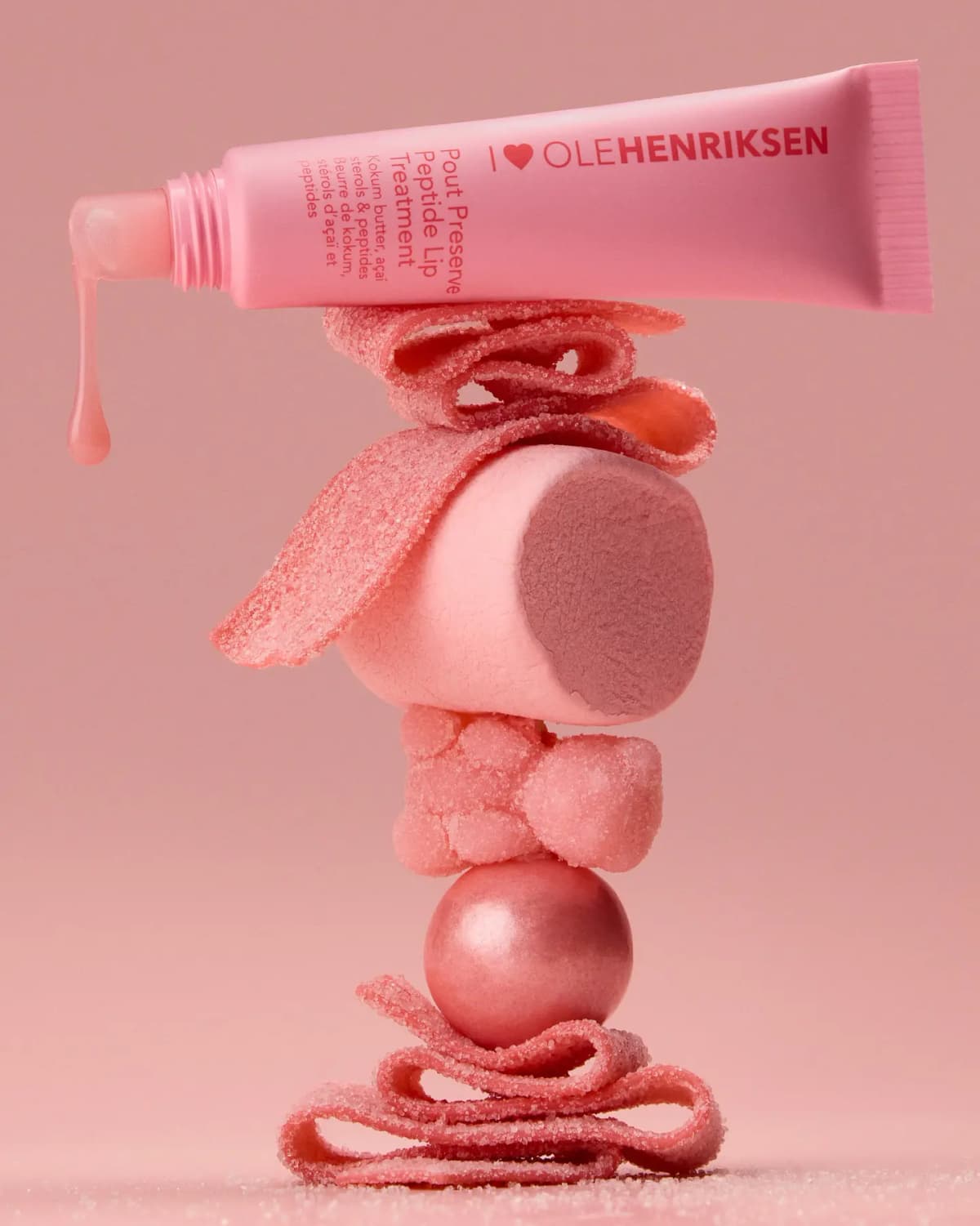 OLEHENRIKSEN Pout Preserve Peptide Lip Treatment tube dripping pink gloss, balanced on stacked pink sugared candies