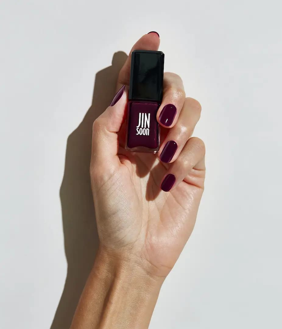 Light-skinned hand with glossy dark plum nails holding a matching JINsoon polish bottle in bright sunlight