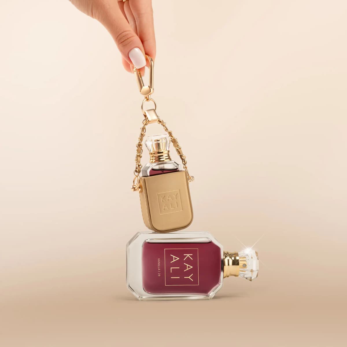 Hand holding a gold clasp with the mini bottle in its chain charm above the 50 ml bottle lying on its side