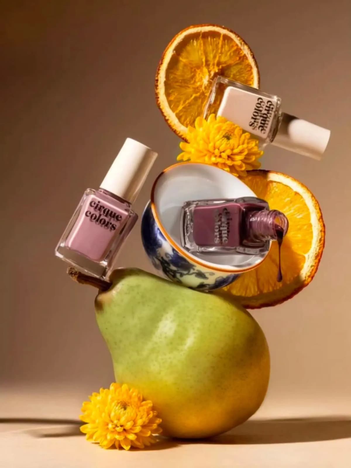 Lilac, plum and cream polish bottles balanced on a pear with dried orange slices and yellow chrysanthemums