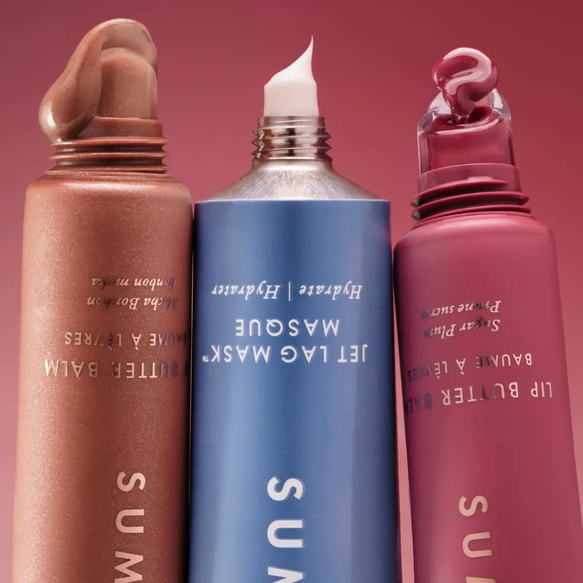 Open Mocha Bonbon balm, Jet Lag Mask and Sugar Plum balm tubes squeezing out product against a pink backdrop