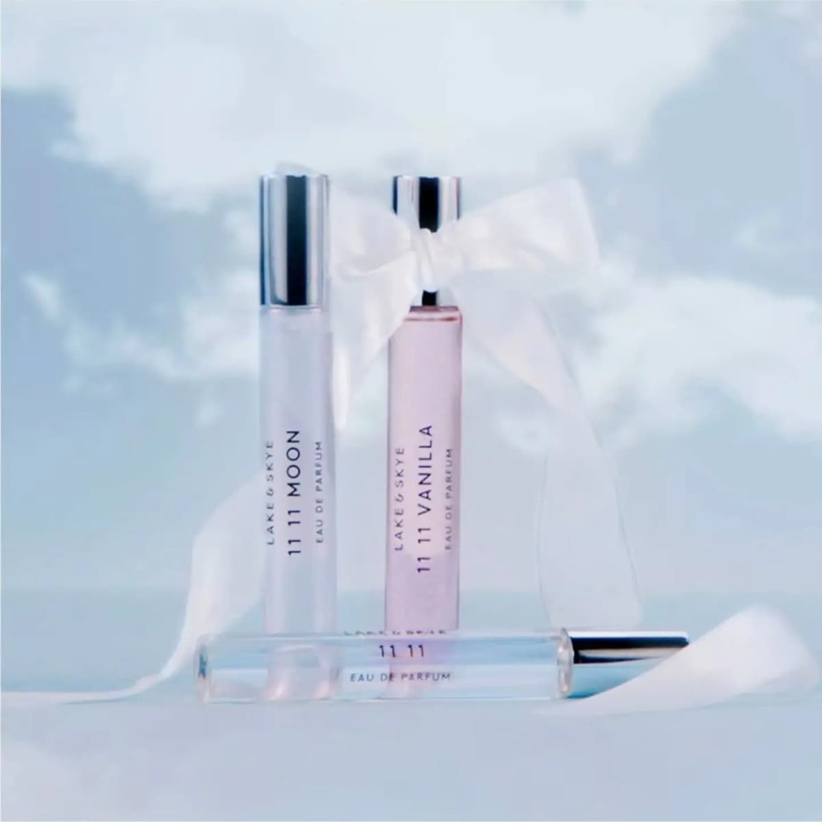 Three Lake & Skye 11 11 travel sprays styled with a white satin ribbon against a soft blue cloudy backdrop.