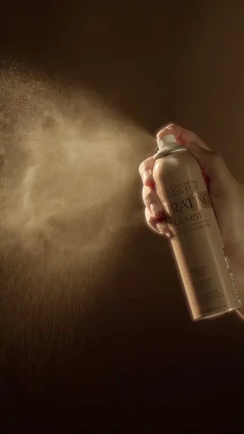 Hand pressing the Hydrating Face Mist nozzle, releasing a fine golden spray cloud against a dark brown background