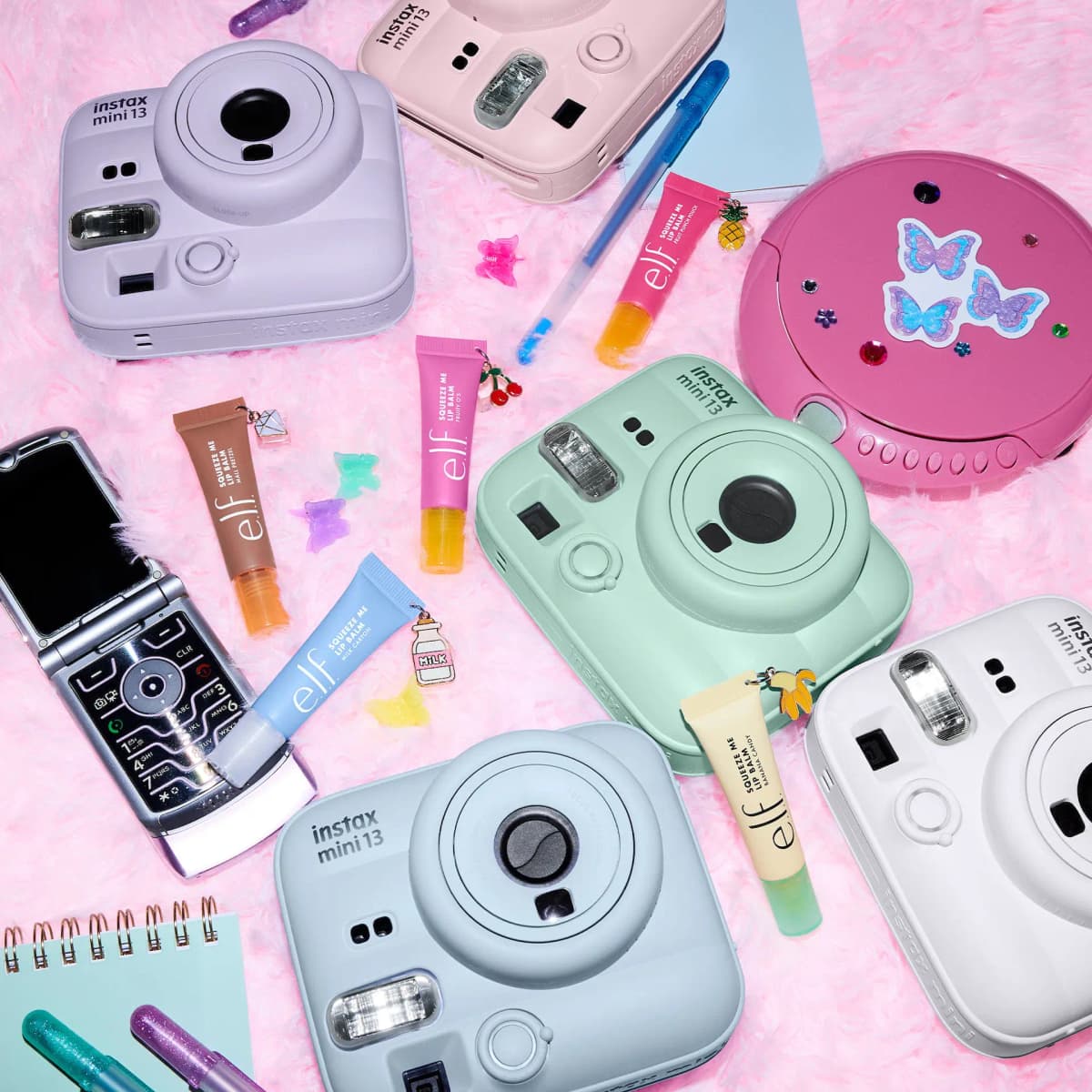 Five lip balm tubes scattered on pink faux fur among pastel instax cameras, a flip phone and a CD player