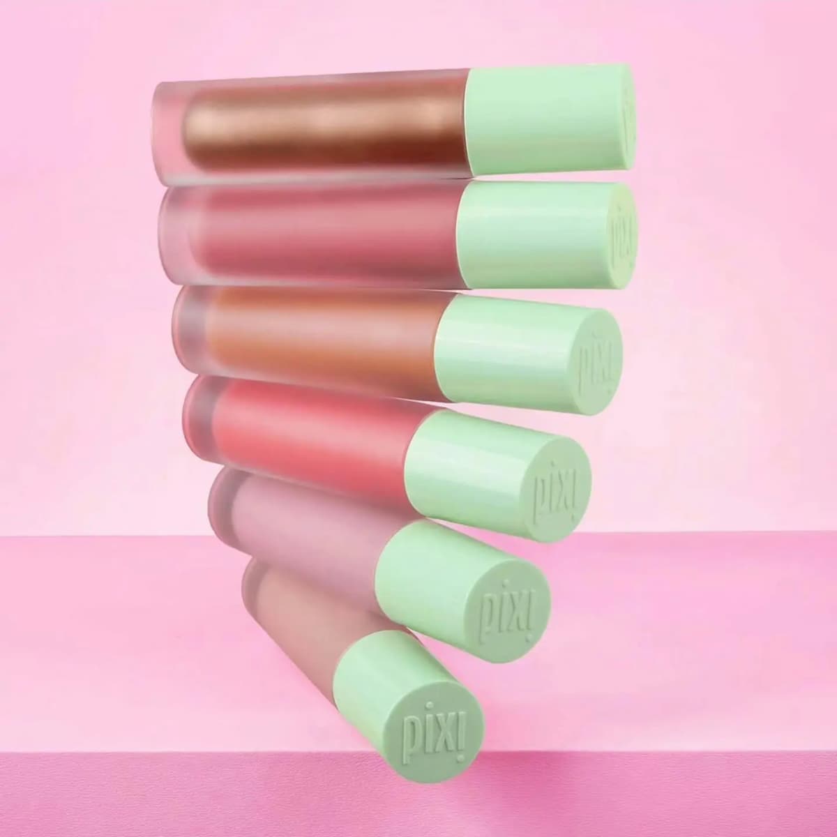 Six frosted blush tubes with mint green Pixi-embossed caps stacked horizontally against a soft pink backdrop.