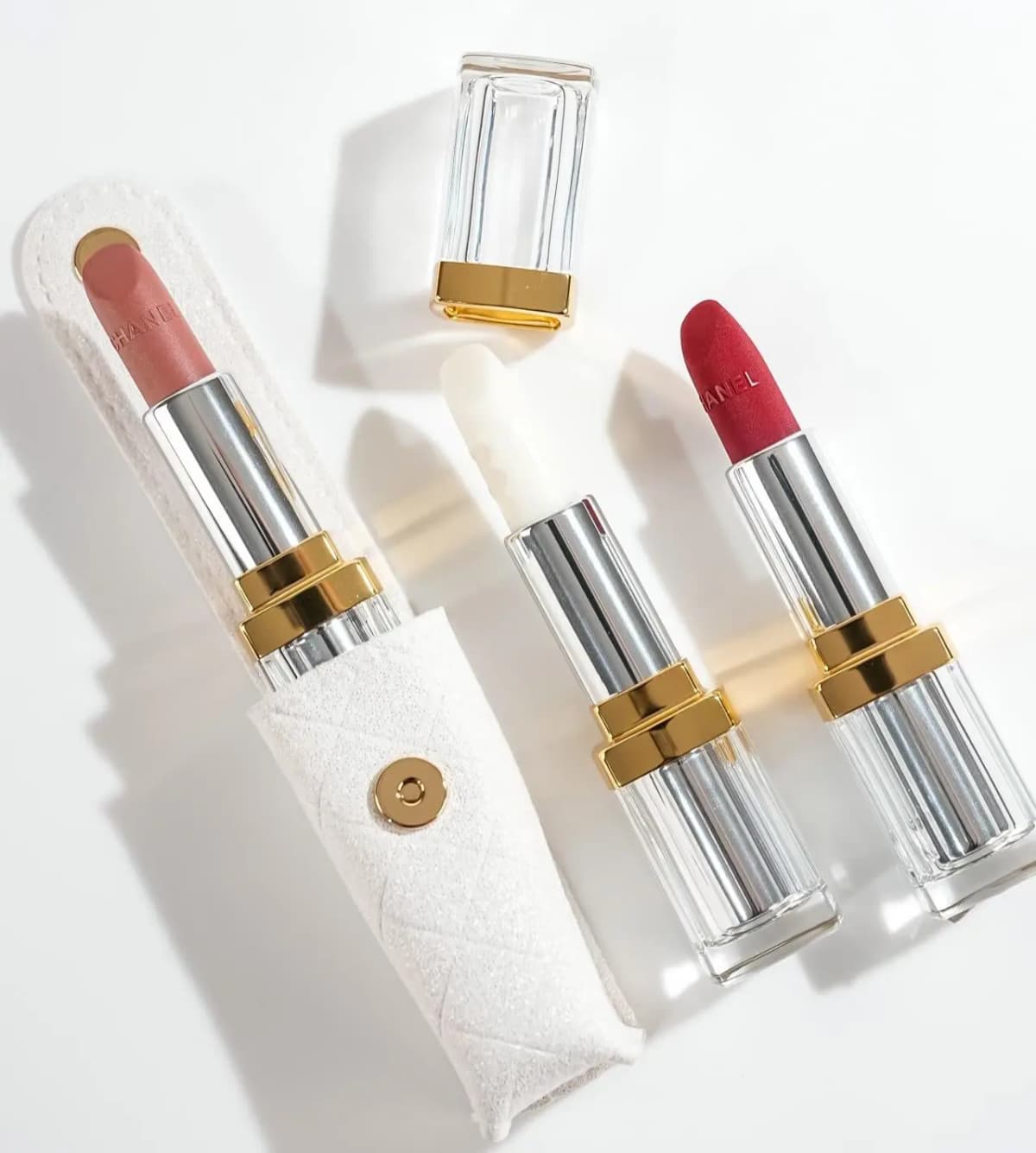 Chanel Holiday 2026 pink, white balm and red lipsticks in clear cases beside a quilted white sleeve