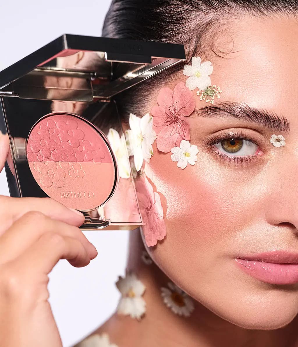 Model with pressed flowers on the face holds the open two-tone pink blush compact beside a rosy cheek