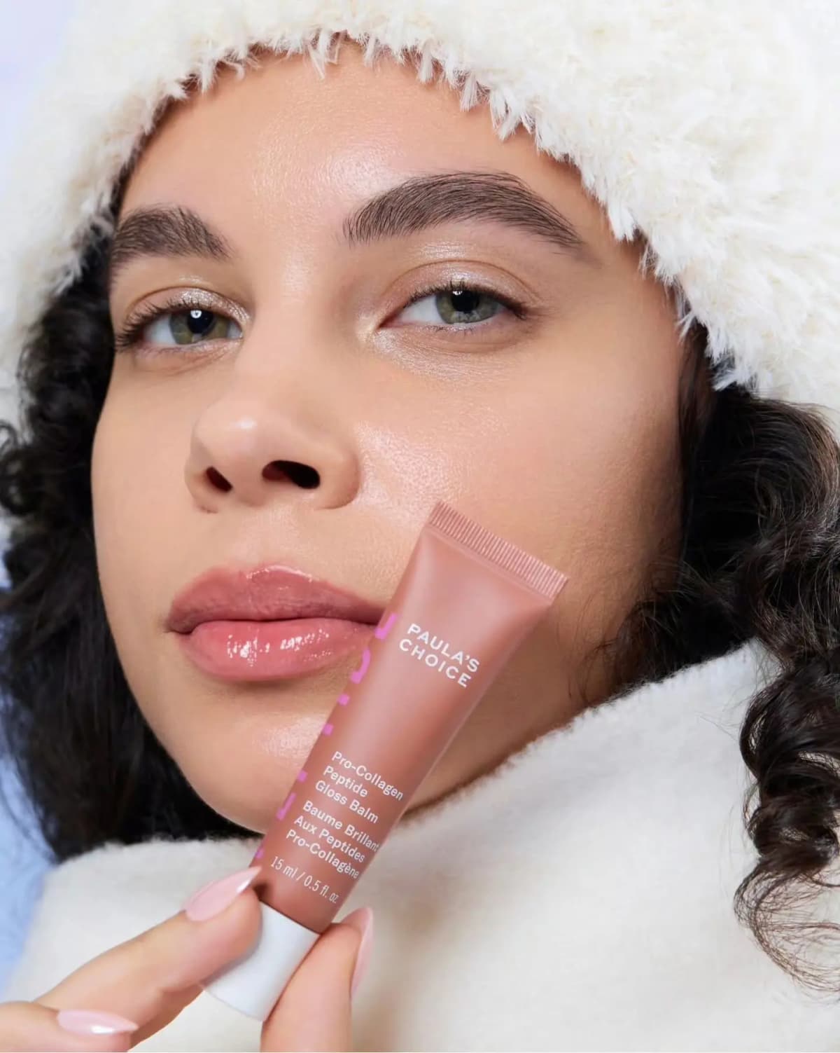 Model in a cream fur hat and coat with glossy nude lips holds the brown gloss balm tube beside her cheek