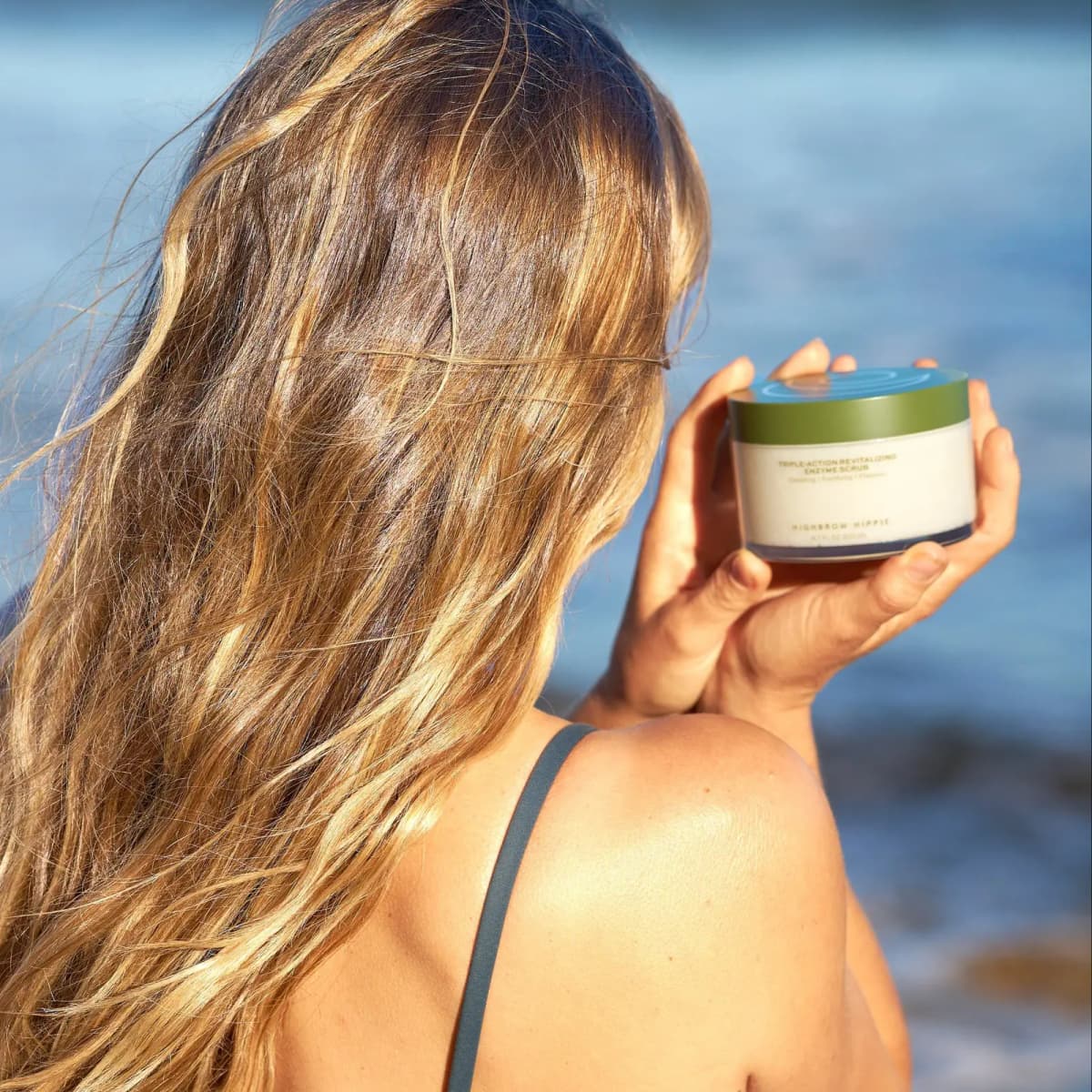Woman with long wavy sun-streaked hair holds the green-lidded scrub jar up by her shoulder against blue sea