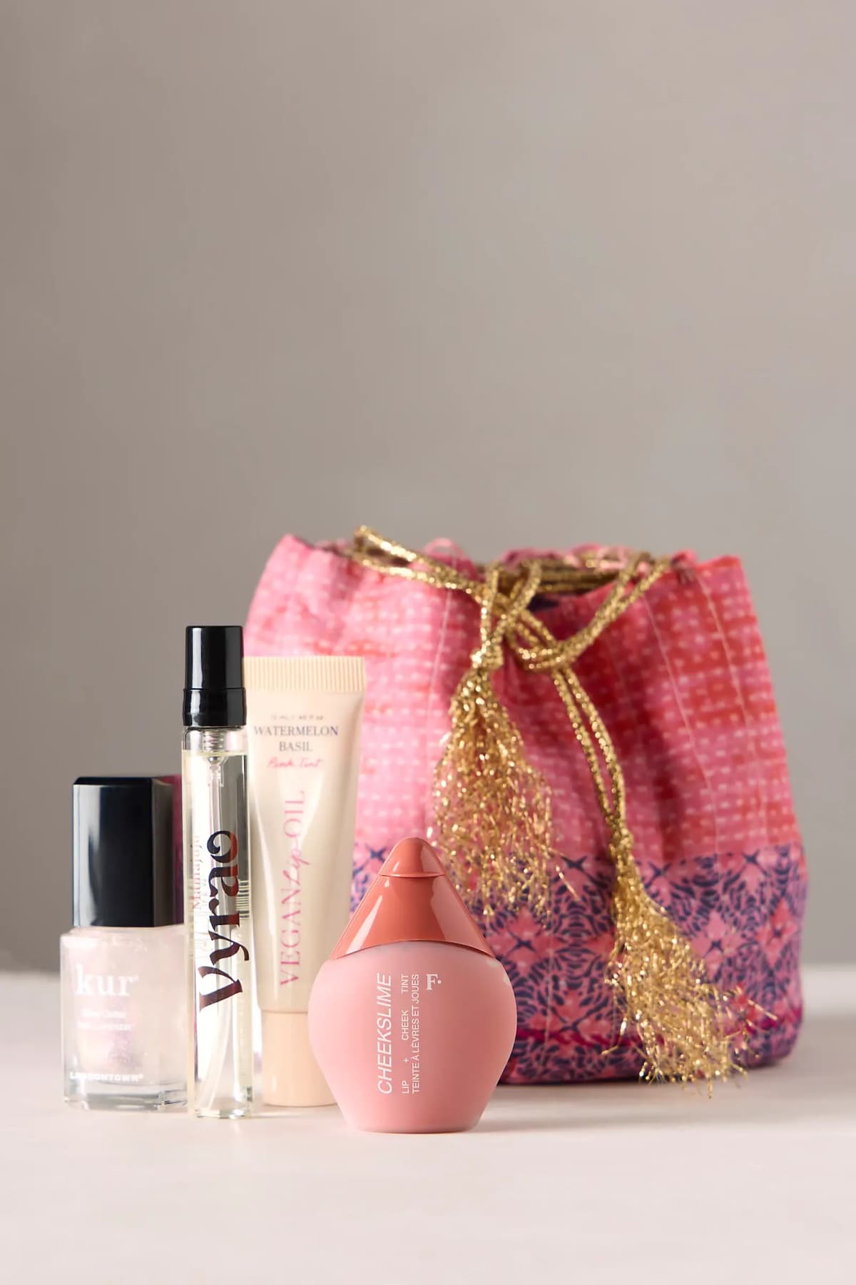 Kur nail polish, Vyrao travel spray, lip oil tube and Cheekslime tint lined up before a pink quilted drawstring bag
