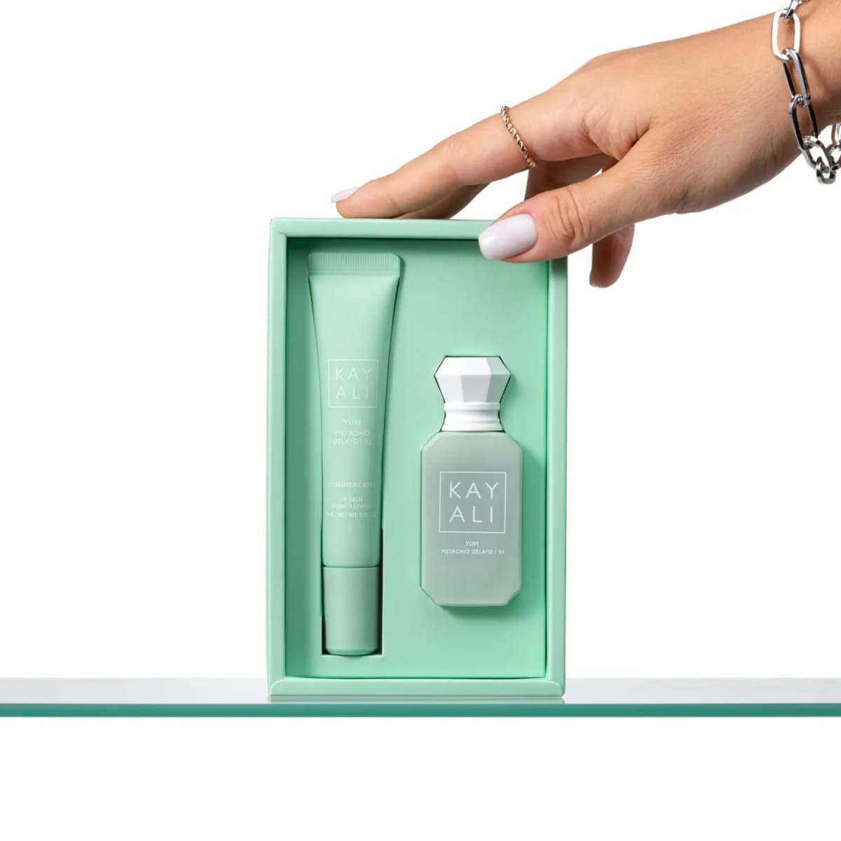 Hand with rings and a chain bracelet holding the open mint box showing the lip balm and pistachio perfume