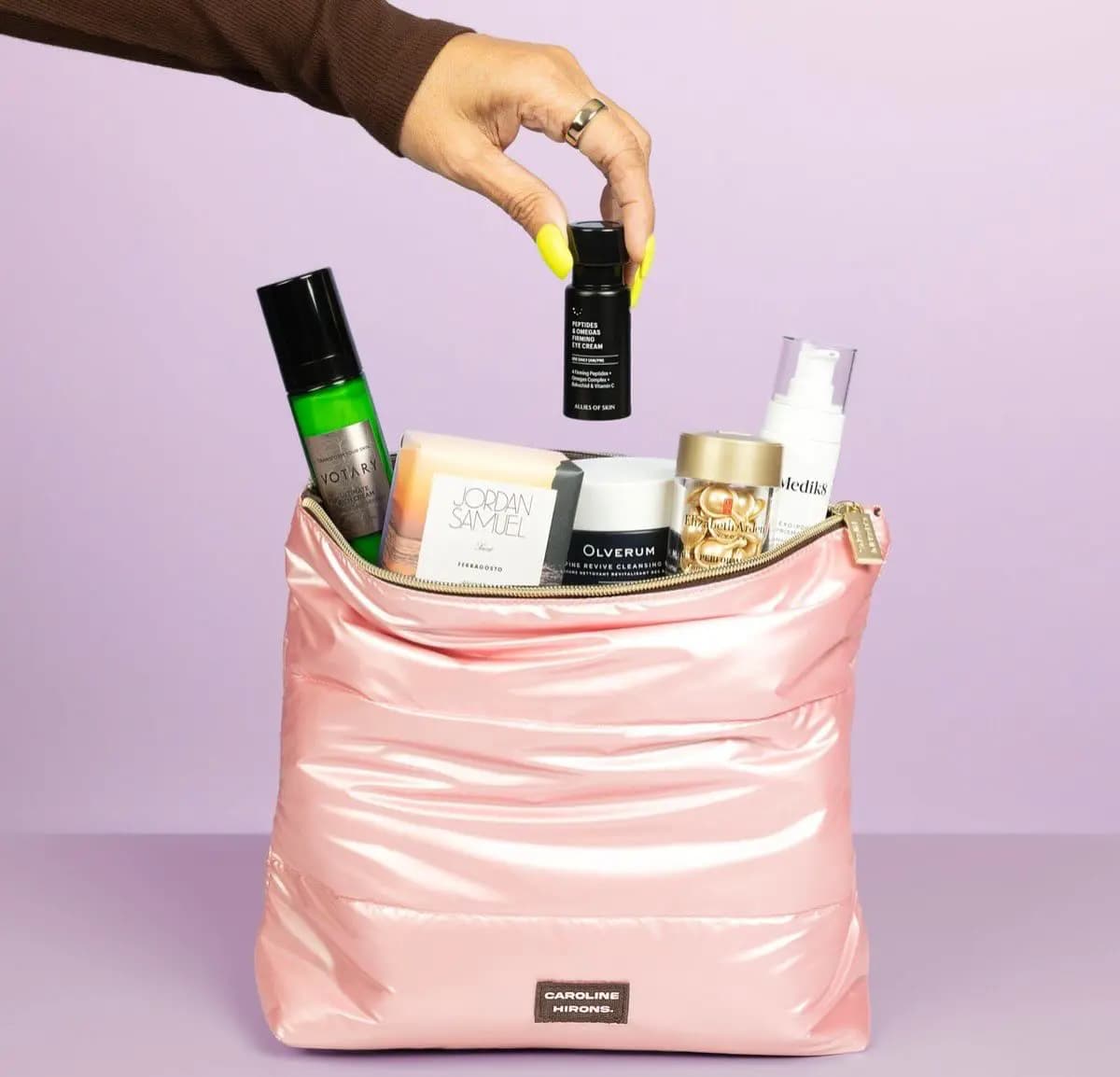 Hand with yellow nails drops Allies of Skin eye cream into a pink puffer Caroline Hirons bag packed with skincare