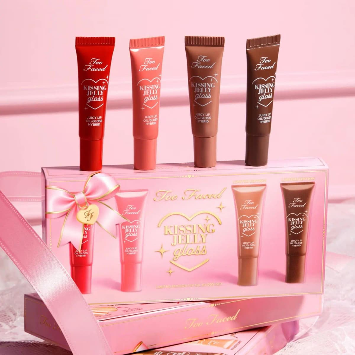Four Too Faced Kissing Jelly Gloss tubes in red, coral, nude and brown atop a pink limited edition gift box with a bow