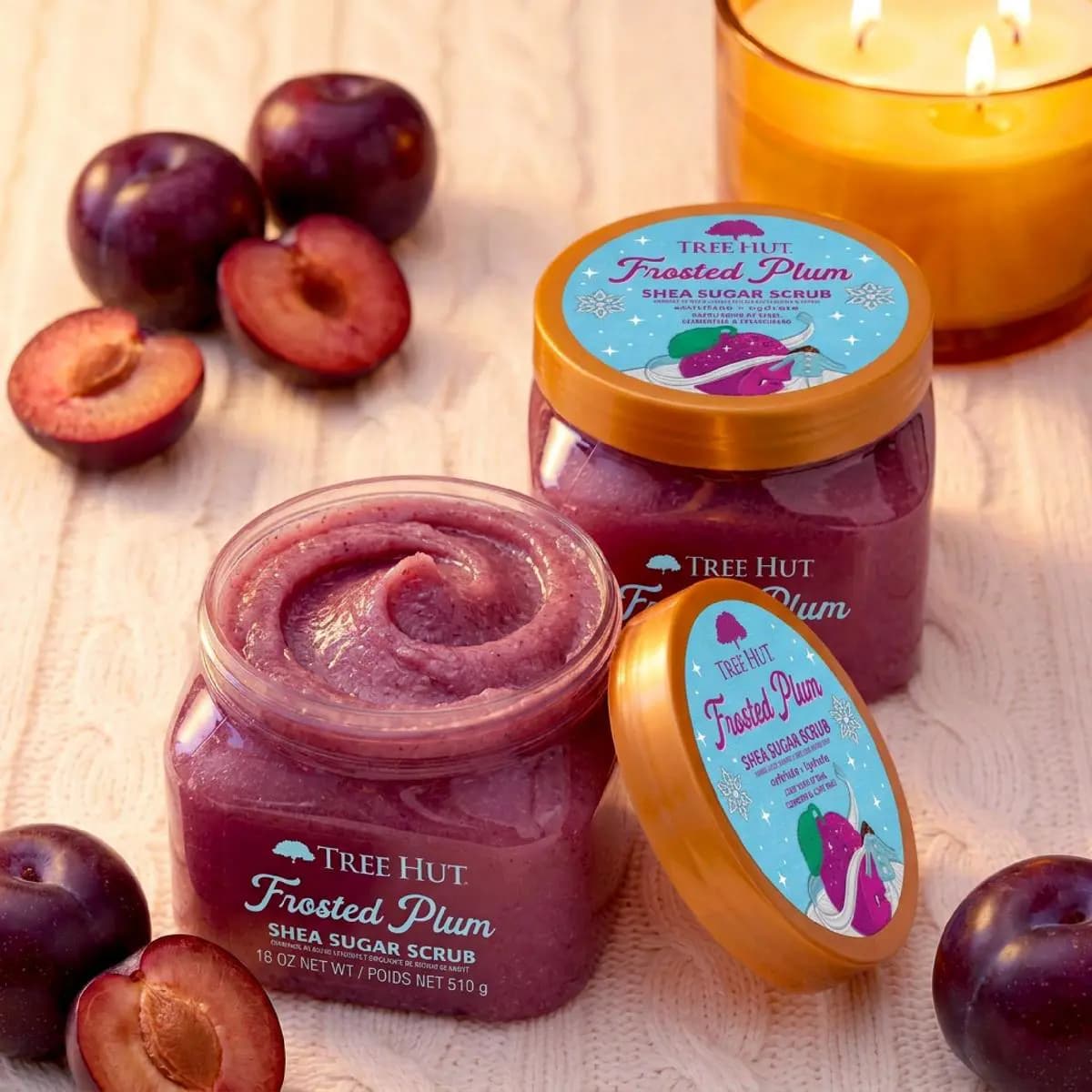 Two Frosted Plum Shea Sugar Scrub jars in deep purple with fresh plums and a lit candle on knit fabric