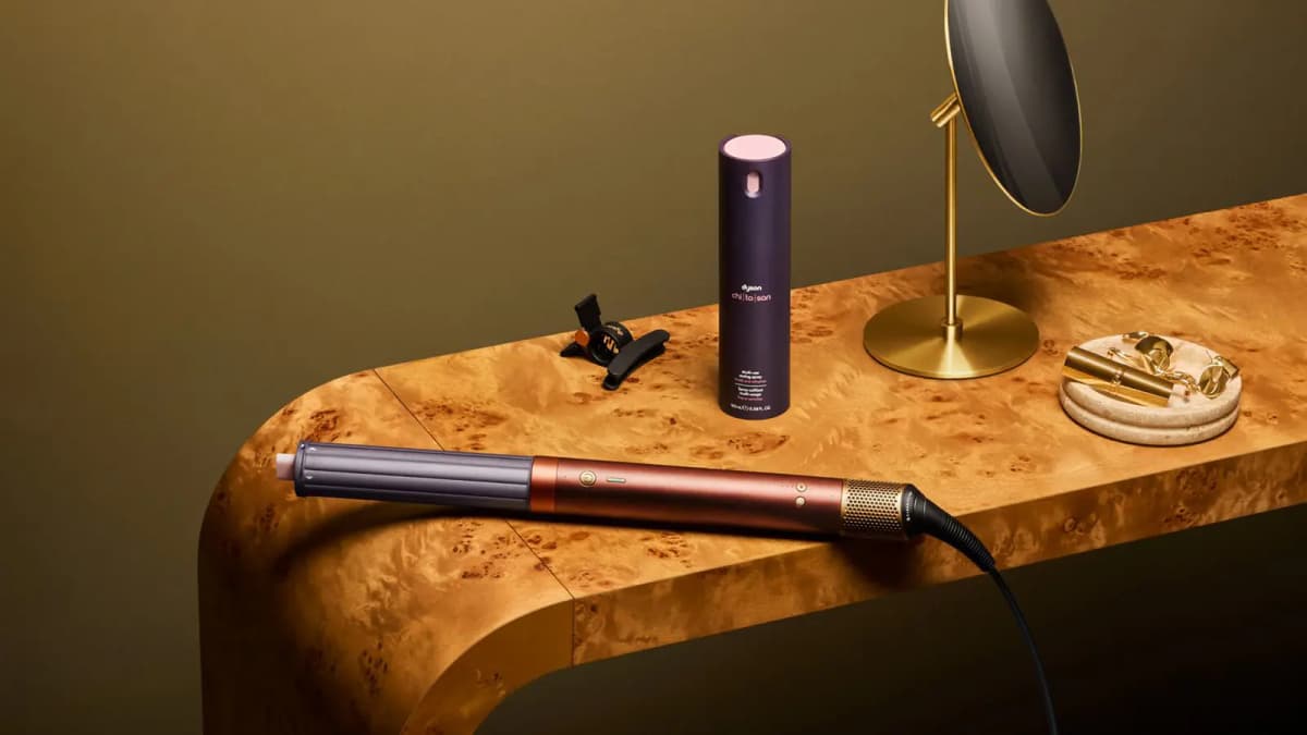 Copper multi-styler with plum barrel on a burl wood console beside a Dyson chi|to|san spray, hair clip and gold mirror
