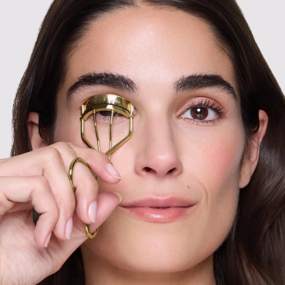 Model curling the upper lashes with a gold eyelash curler held up to one eye