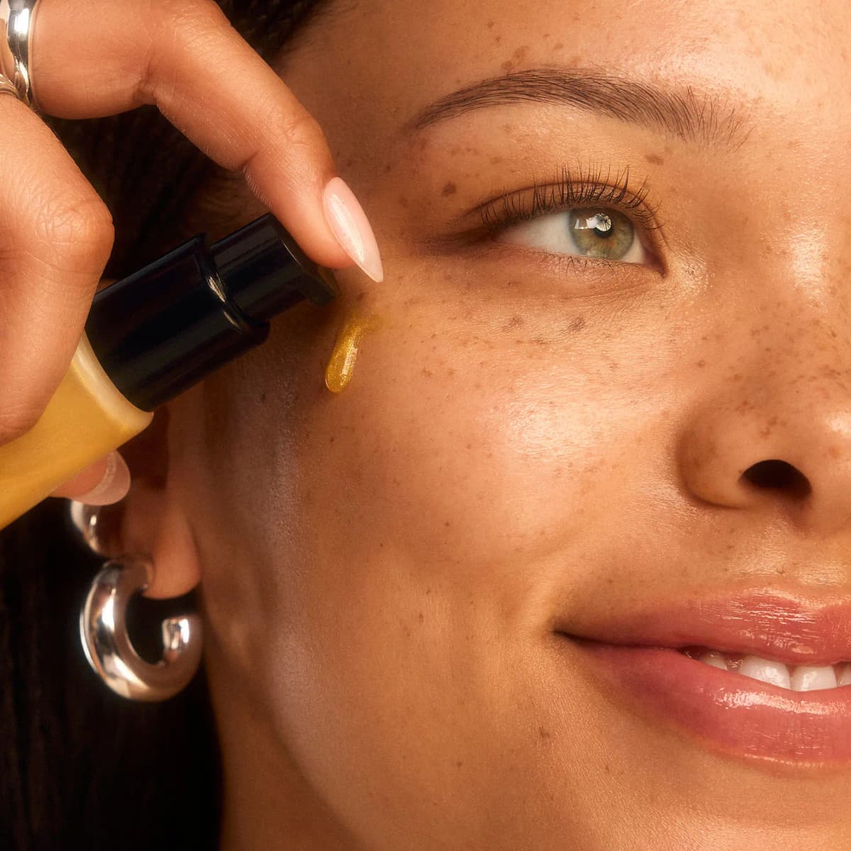 Hand with almond nails pumping golden shimmer primer onto a freckled model's cheekbone.