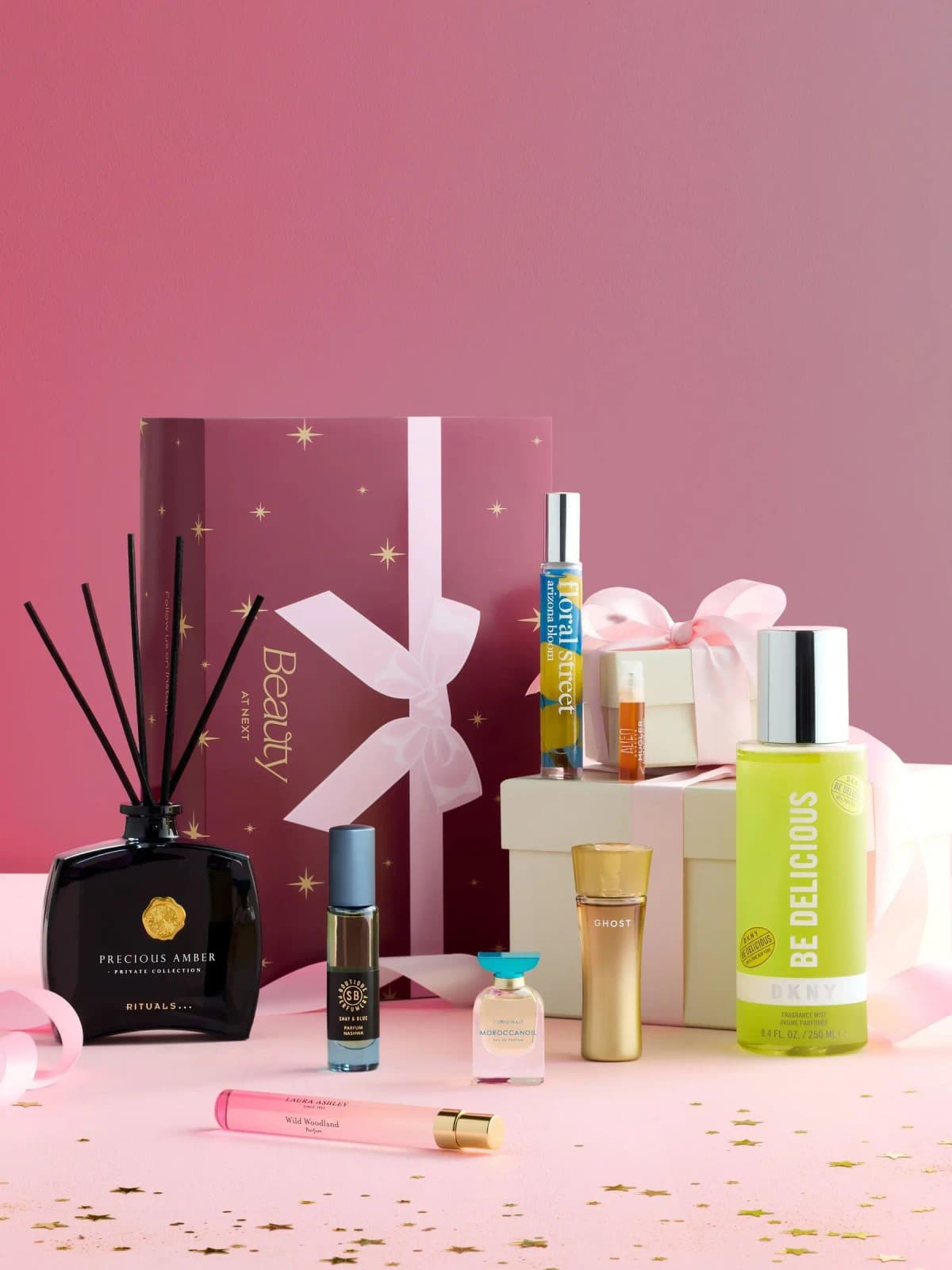 Beauty at Next Wild Botanicals edit: Rituals diffuser, DKNY mist and six perfumes before a plum star-print box on pink.