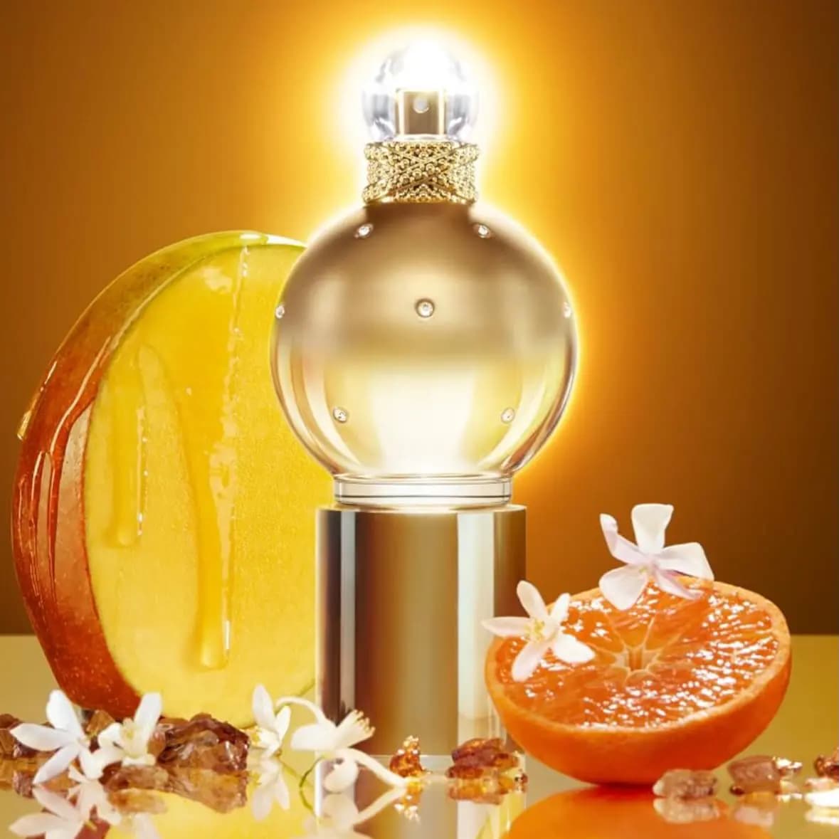 Spherical gold perfume bottle on a gold pedestal surrounded by mango slice, orange half, jasmine blossoms and amber crystals.