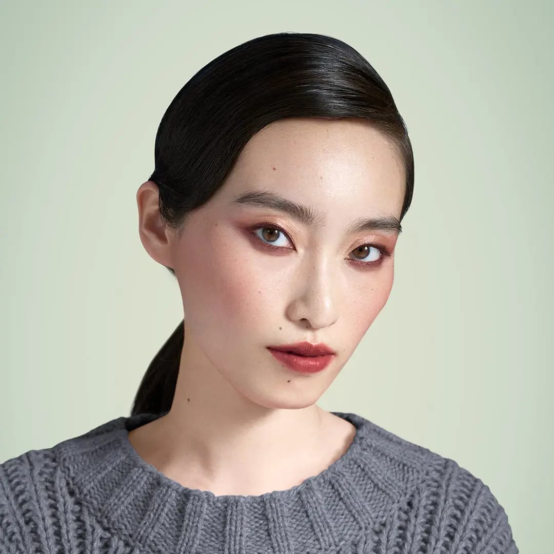 Model with sleek ponytail wearing copper and burgundy eye look with red-brown lips against a pale green backdrop
