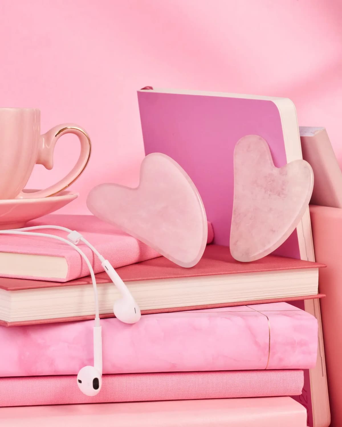 Two pink heart-shaped gua sha stones leaning on a stack of pink books with earphones and a teacup