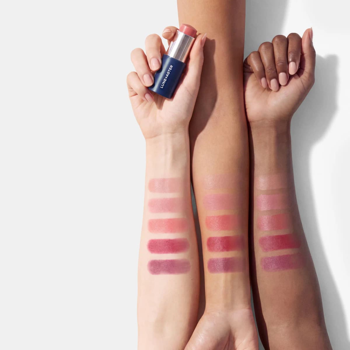 Five cream stick shades swatched on three forearms of light, medium and deep skin tones, one hand holding the stick