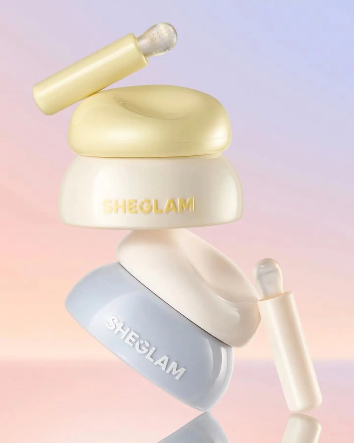 Cream-and-yellow lip scrub jar balanced on a blue lip mask jar, each with an applicator, on a pink-lilac gradient