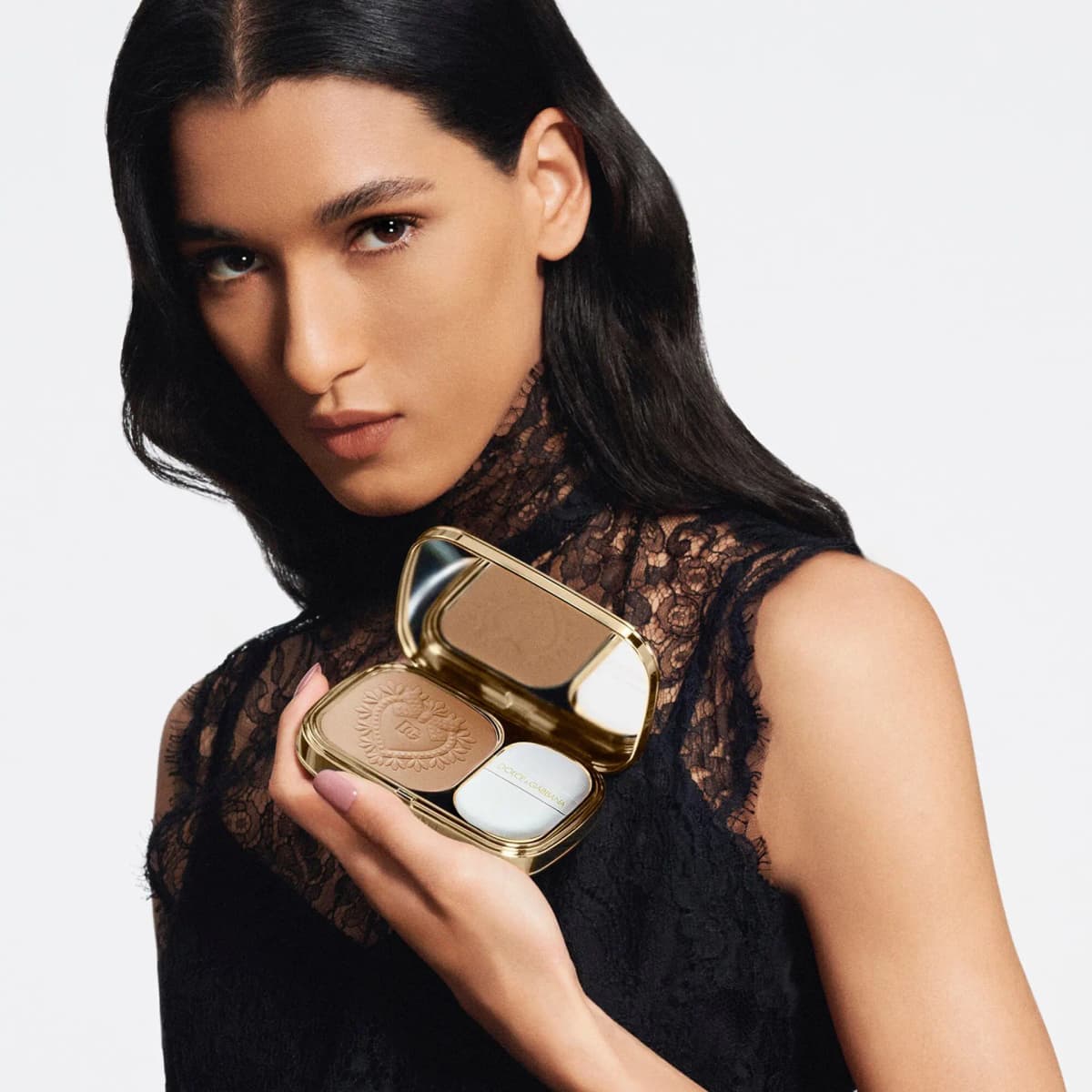 Dark-haired model in a black lace top holds an open gold compact with tan powder, a mirror and a white sponge