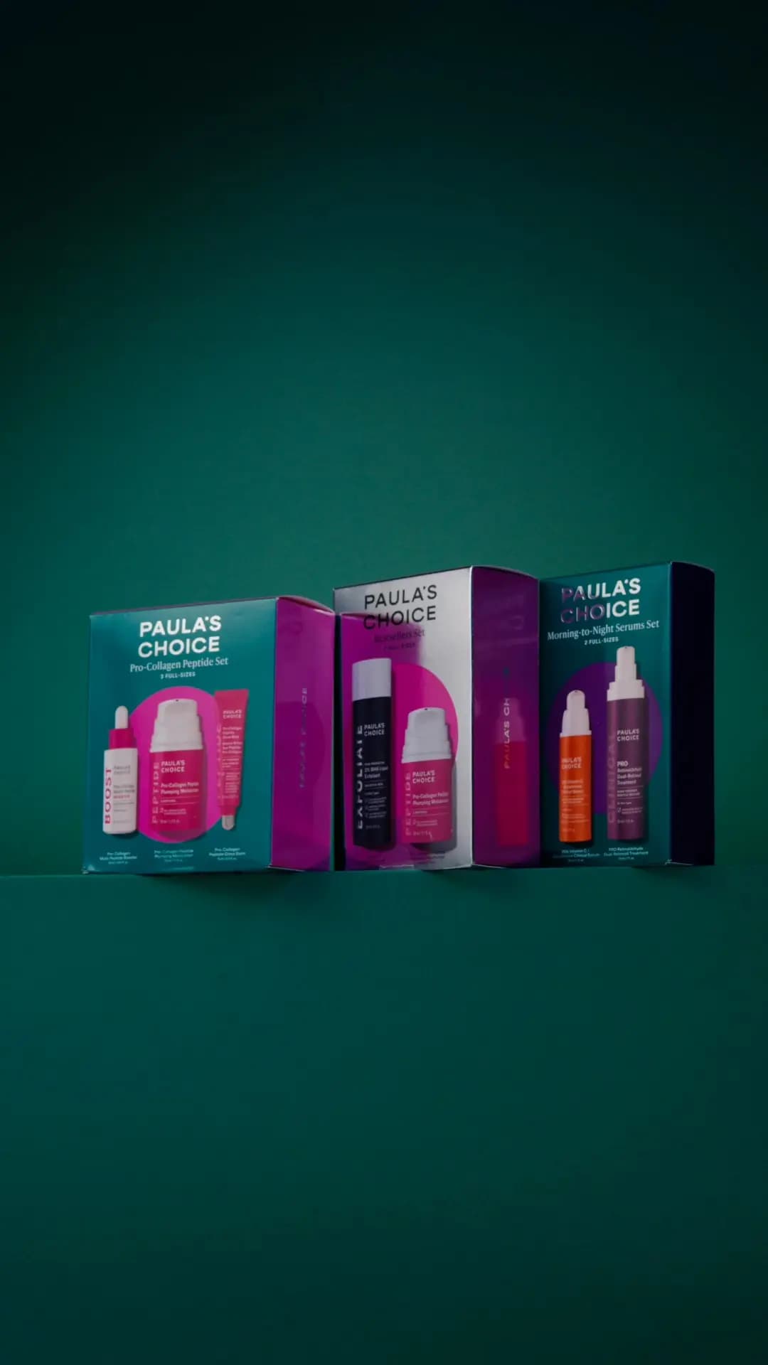 Three Paula's Choice holiday kit boxes in teal, silver and magenta lined up on a ledge against a dark green backdrop.