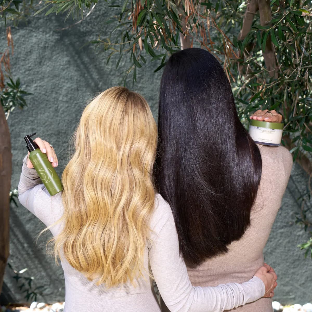 Two women from behind, blonde waves and sleek black hair, holding a green spray bottle and the scrub jar