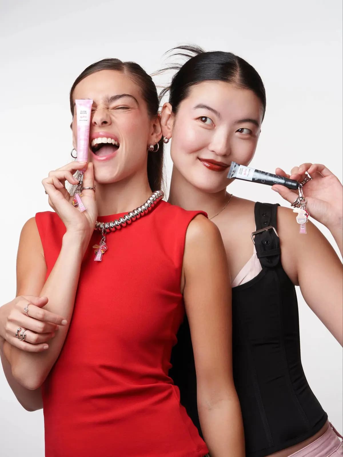 Glow Recipe x Buldak Glass Balm Duo tubes, pink and black, held by two models in red and black tops with keychain charms