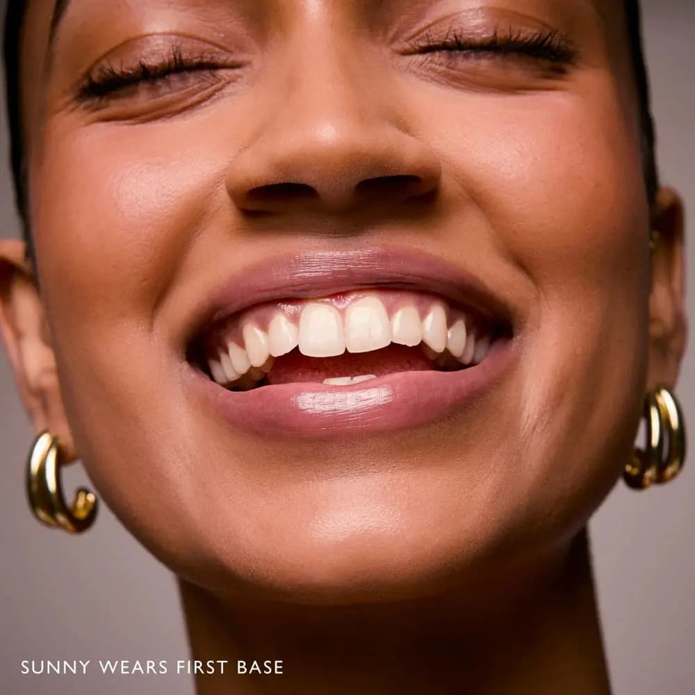 Smiling model Sunny with eyes closed and gold hoops wearing a soft mauve First Base lip
