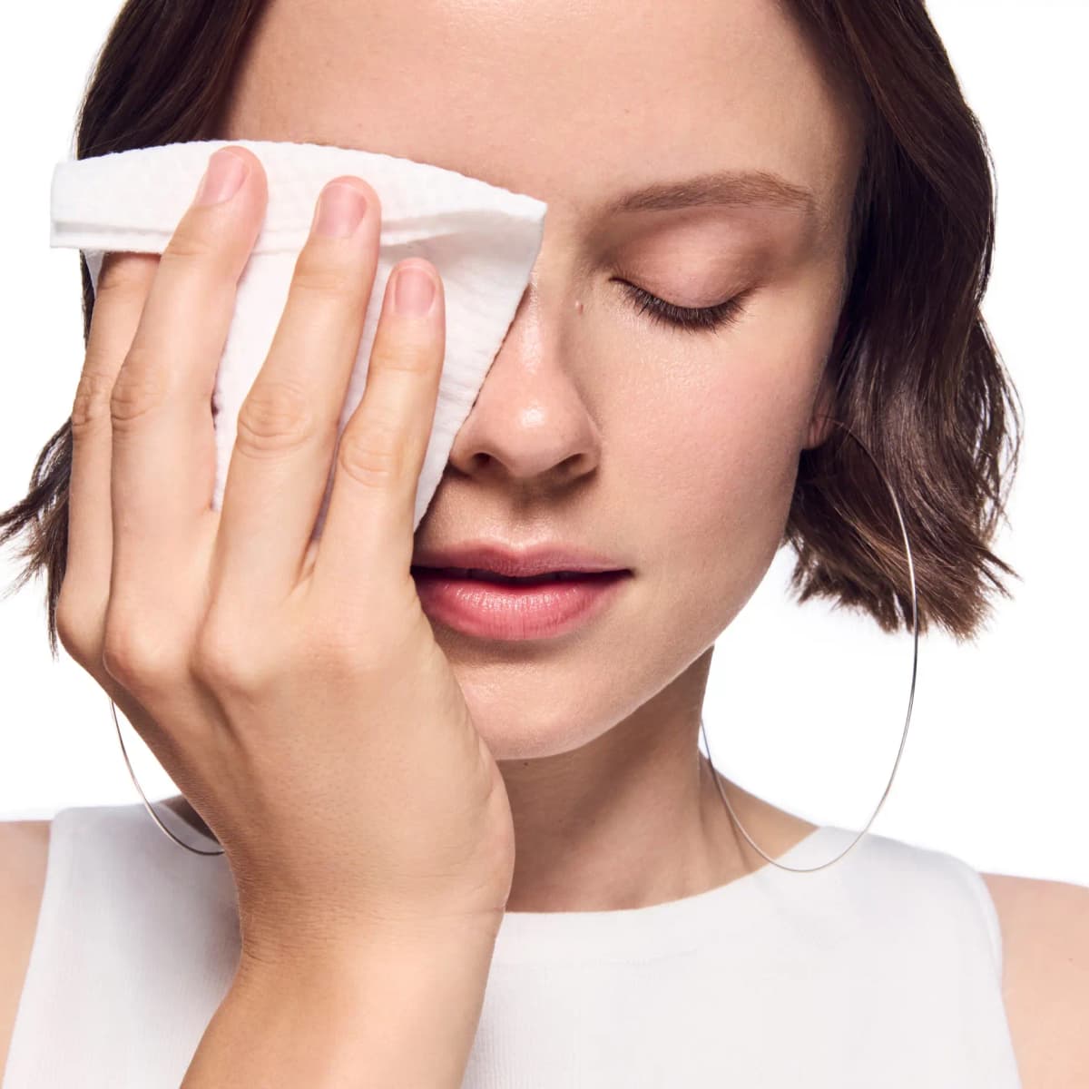 Close-up of a woman with closed eyes gently pressing a white disposable facial towel against her face.