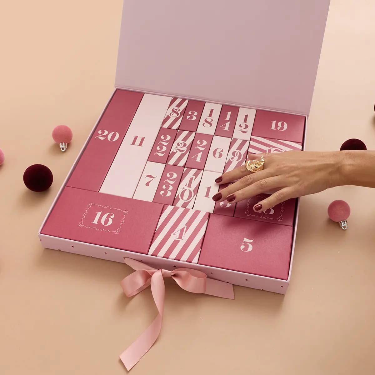 Opened calendar showing pink and striped numbered doors, with a hand in burgundy nail polish reaching in