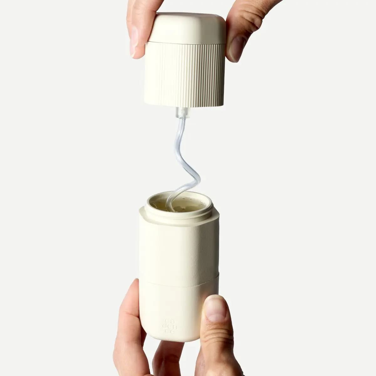 Hands lifting a capped ribbed pump from a cream capsule, its clear dip tube rising out of liquid