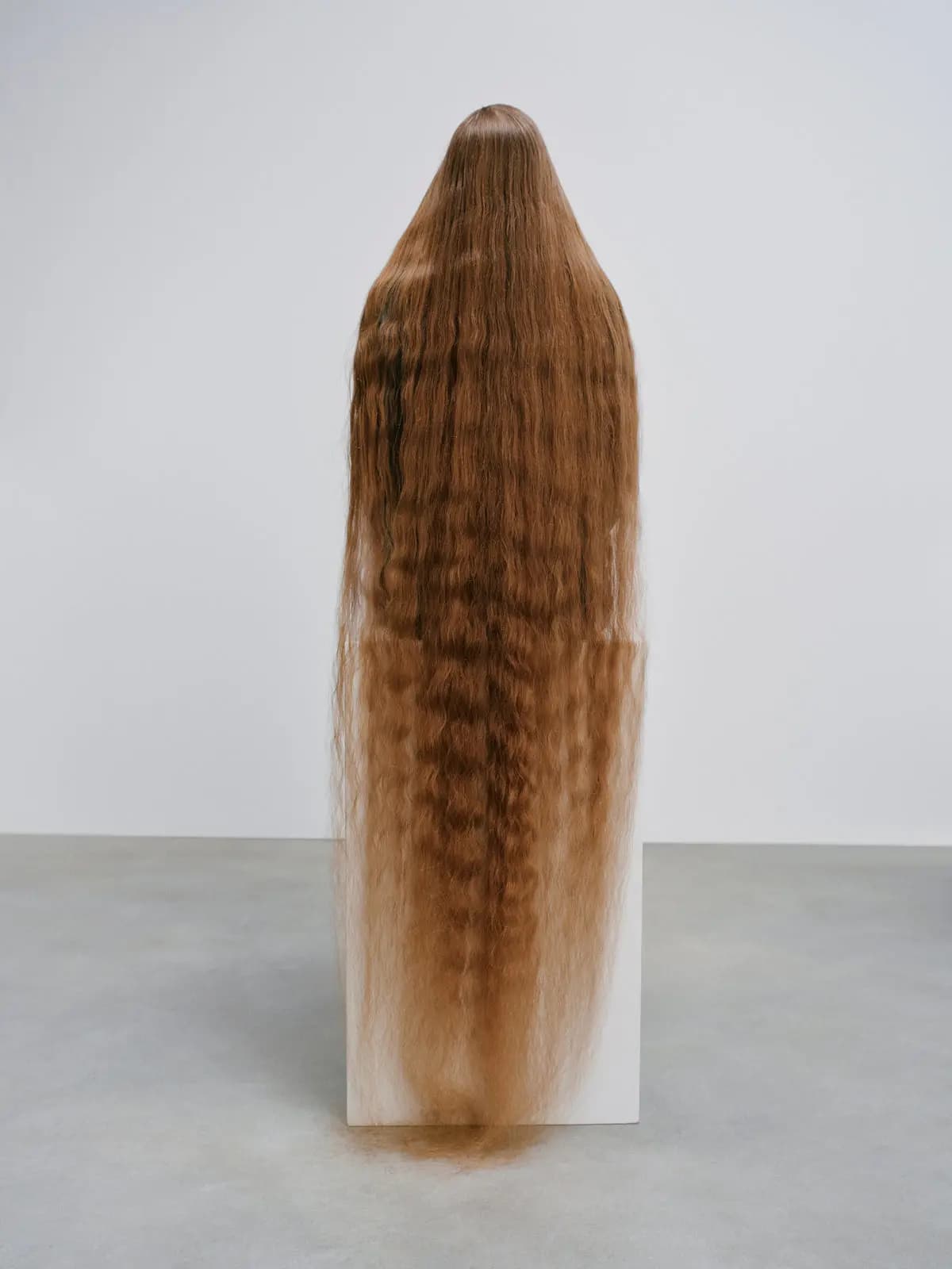 Floor-length crimped auburn hair cascading down the back of a figure seated on a white plinth, face hidden