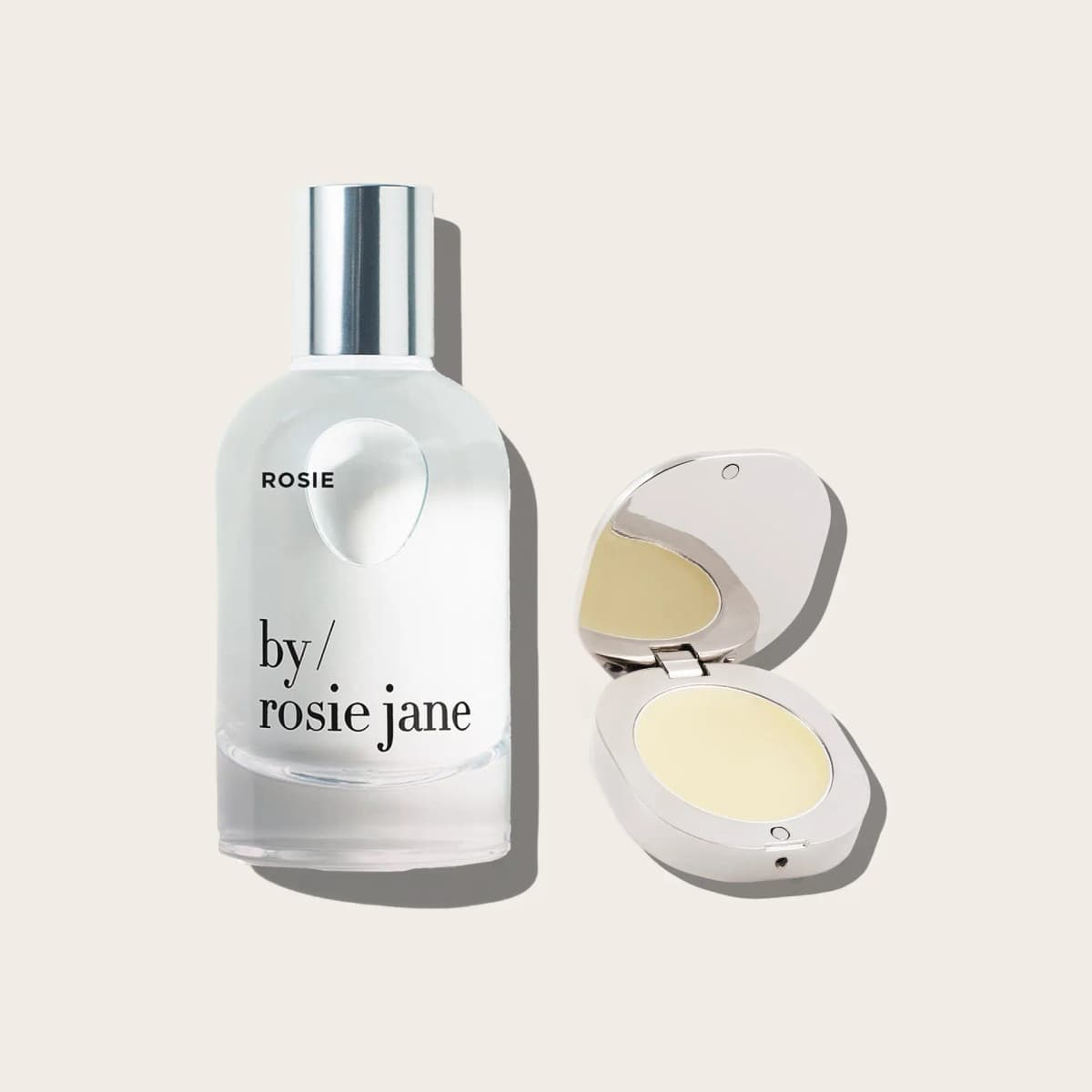 Rosie eau de parfum bottle beside an open silver compact holding pale yellow balm on a cream background