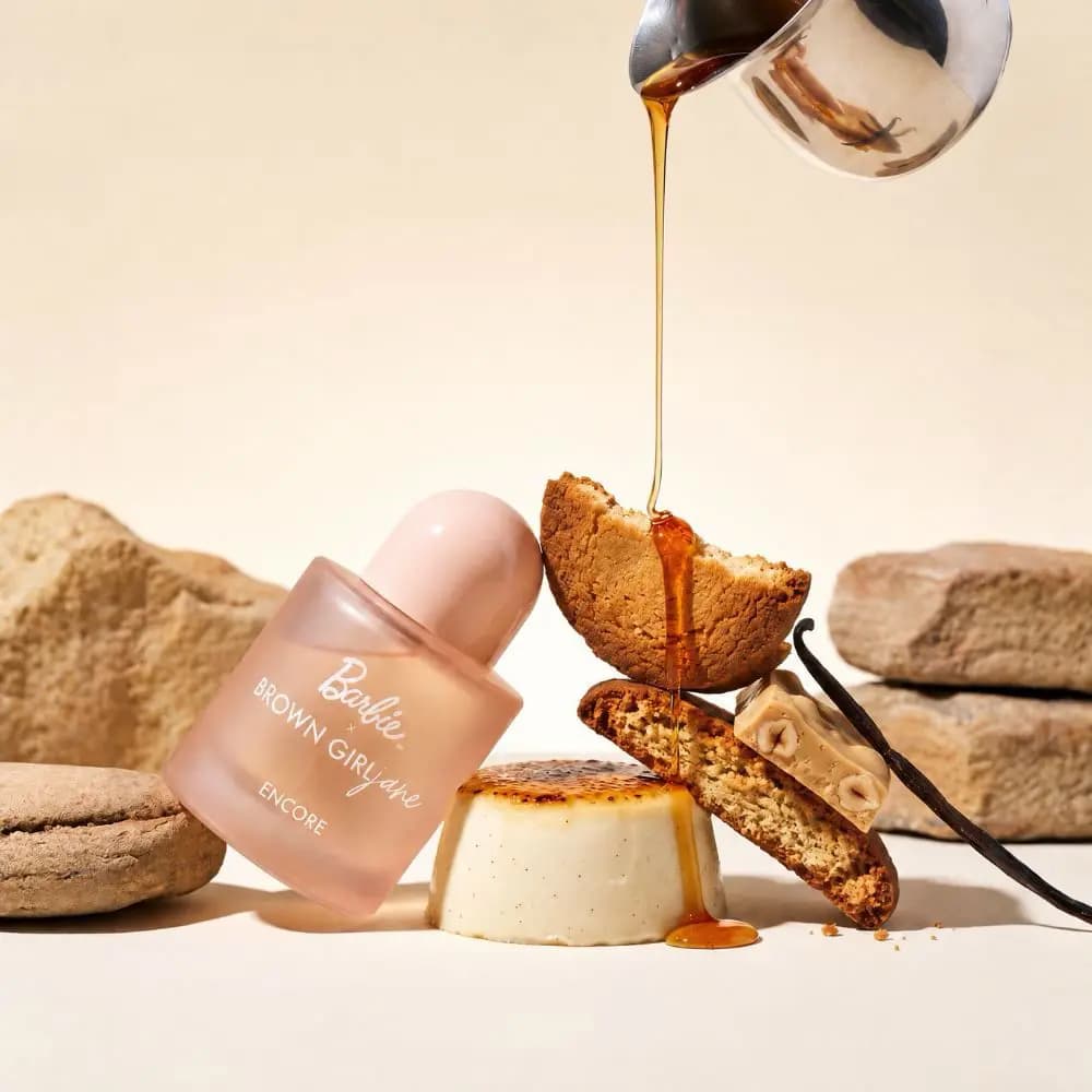Peach perfume bottle leaning on a crème brûlée beside cookies, nougat and vanilla as syrup pours from a silver jug