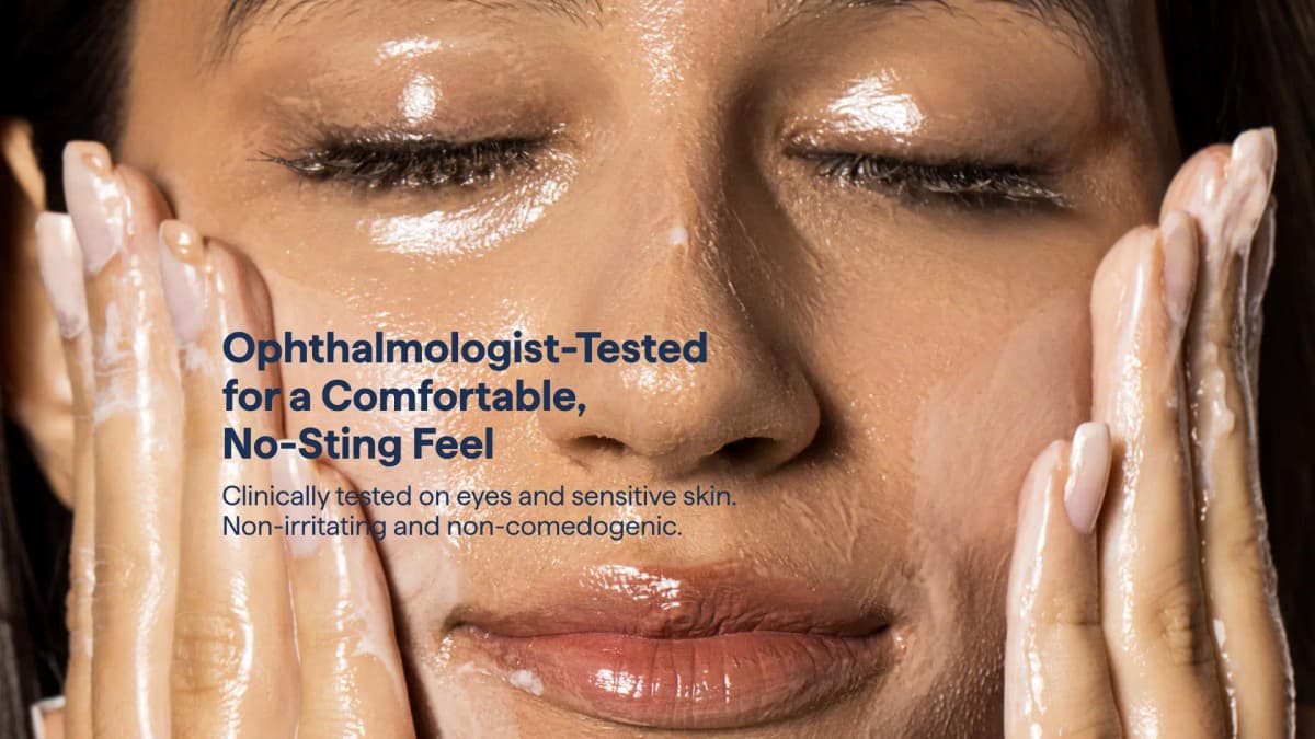 Close-up of a woman with closed eyes massaging glossy, milky oil into her face under ophthalmologist-tested text