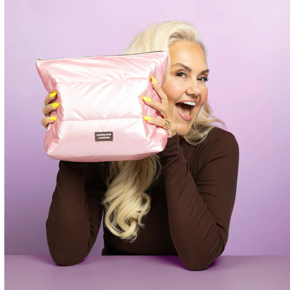 Laughing blonde woman with yellow nails holds a glossy pink quilted Caroline Hirons bag up beside her face