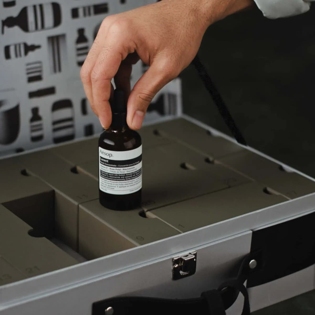 Hand placing an amber Aesop Lucent Facial Concentrate dropper bottle onto the green numbered drawers of the open calendar.
