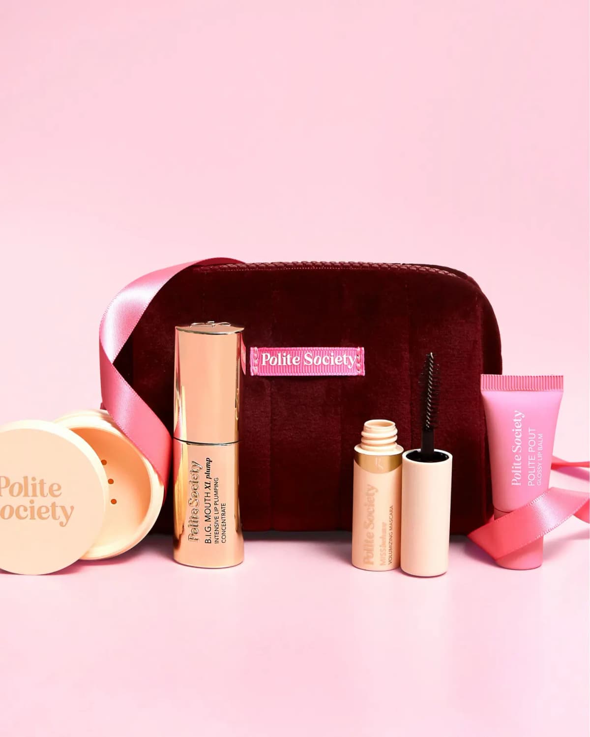 Burgundy velvet Polite Society mini bag with pink label surrounded by gold lip plumper, mascara, pink lip balm and powder jar