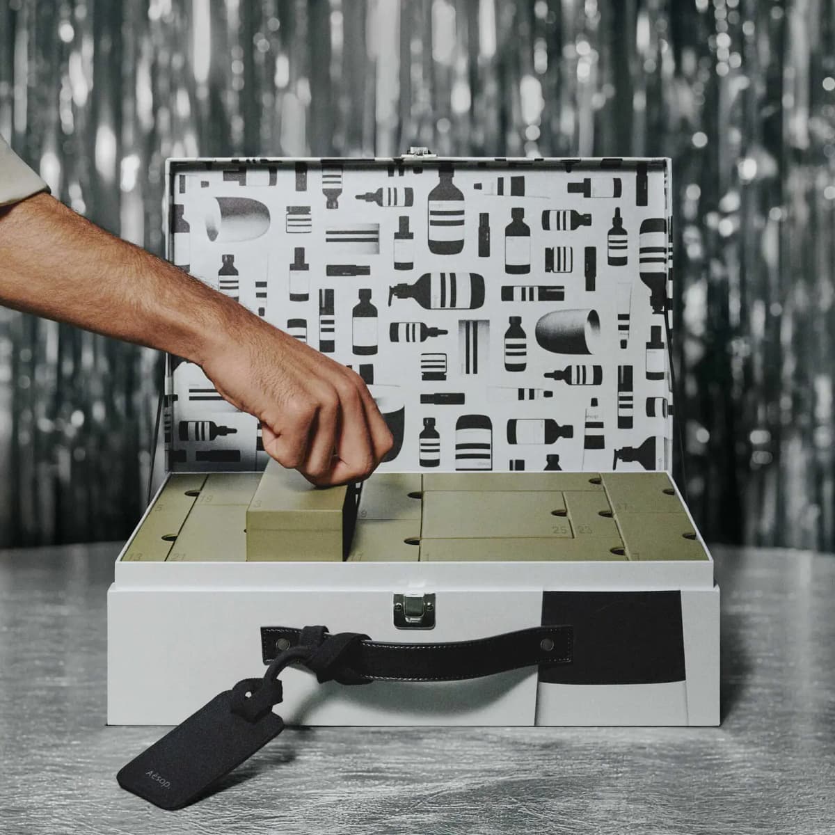 Hand lifting a numbered drawer from the open Aesop Valise calendar against a shimmering silver backdrop.