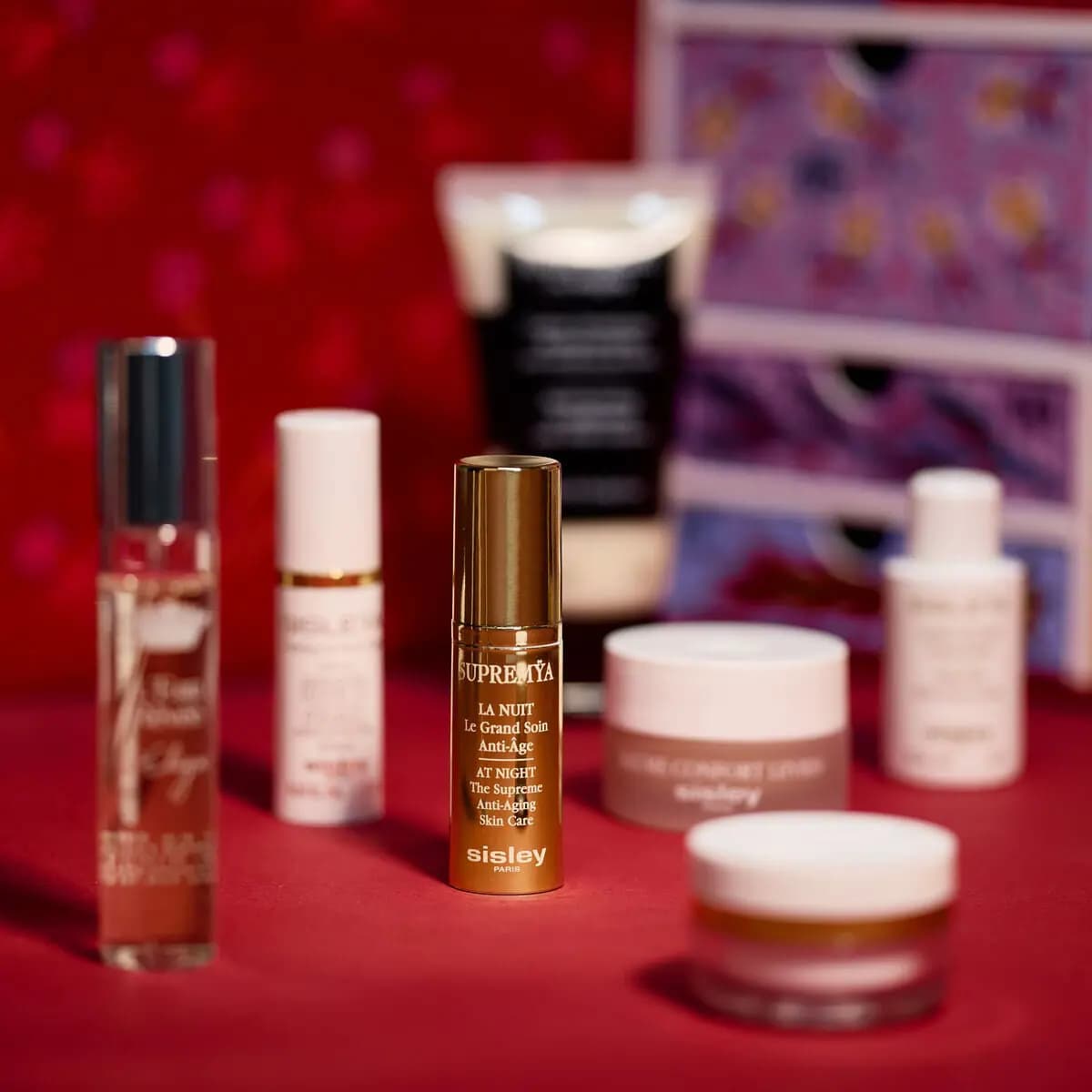 Gold Supremÿa La Nuit anti-aging skincare bottle in focus beside blurred white jars, tubes and a spray on red