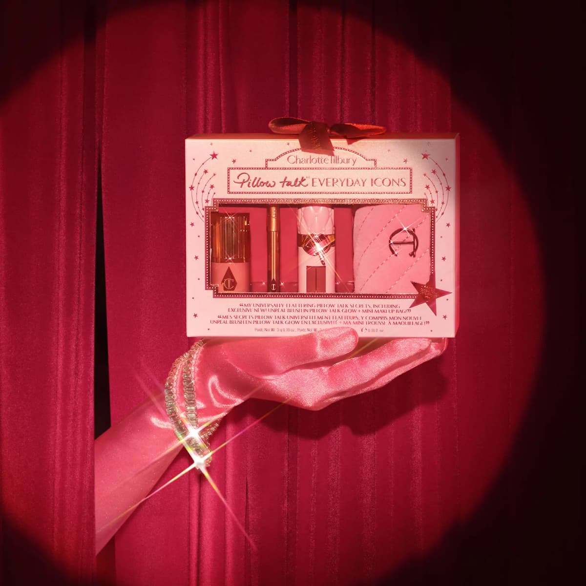 Gloved hand in pink satin holds the gift box under a spotlight against a red velvet stage curtain