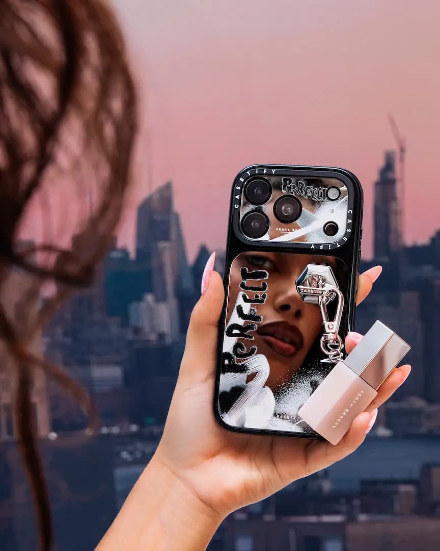 Hand holds a black mirror case with "Perfect" lettering, hexagon charm and lip gloss clipped on, city skyline behind