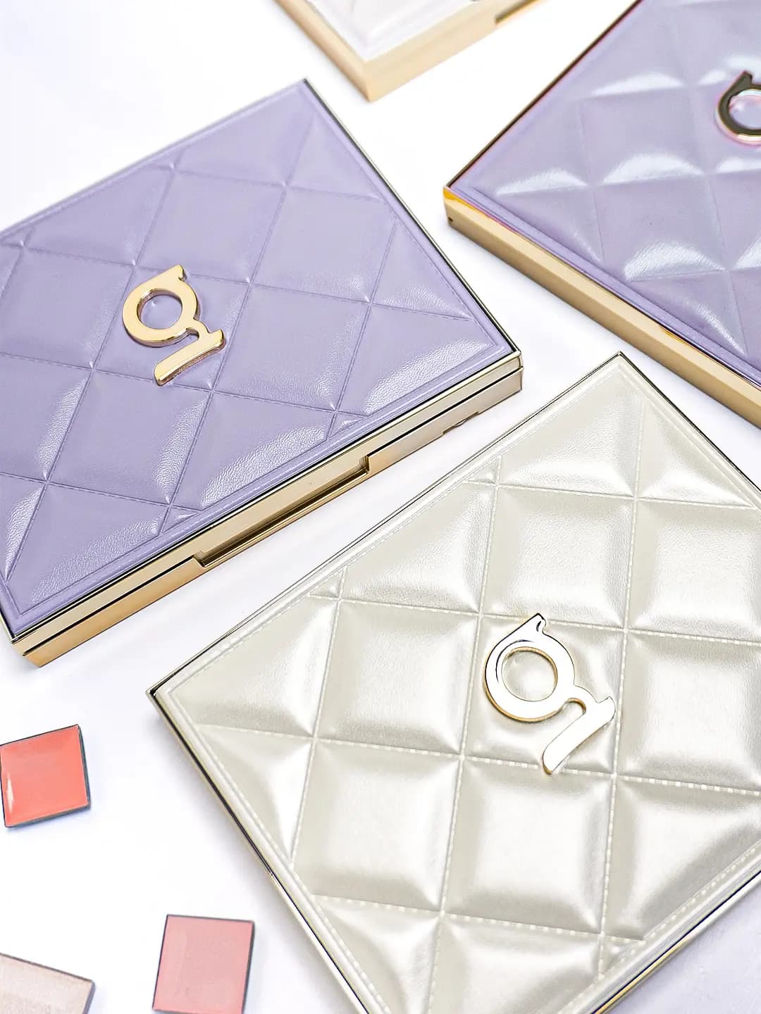 Lavender and pearly ivory quilted puffy palette cases with gold frames and gold g logos, beside loose blush pans