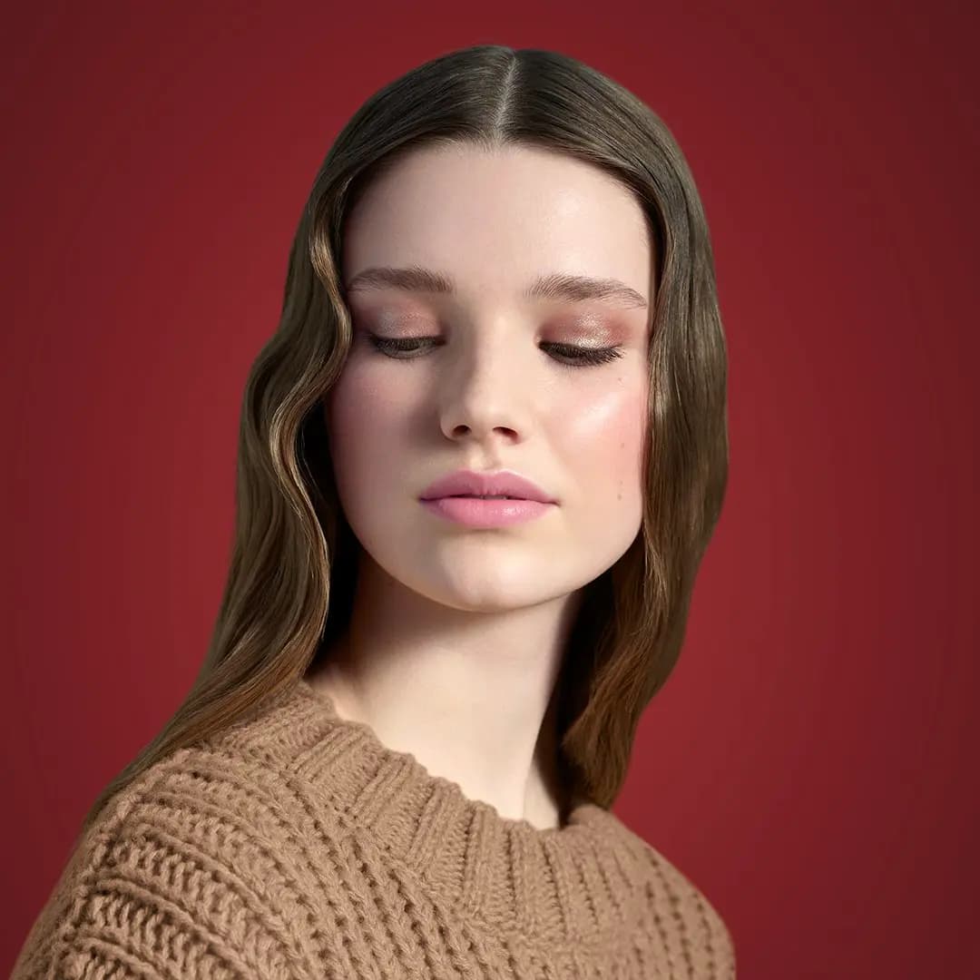 Model with wavy hair and closed eyes wearing rosy shimmer lids and pink lips against a red backdrop