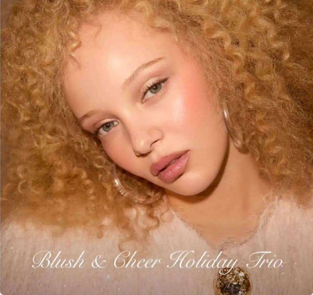 Model with golden curly hair, rosy flushed cheeks and pink lips under "Blush & Cheer Holiday Trio" script text